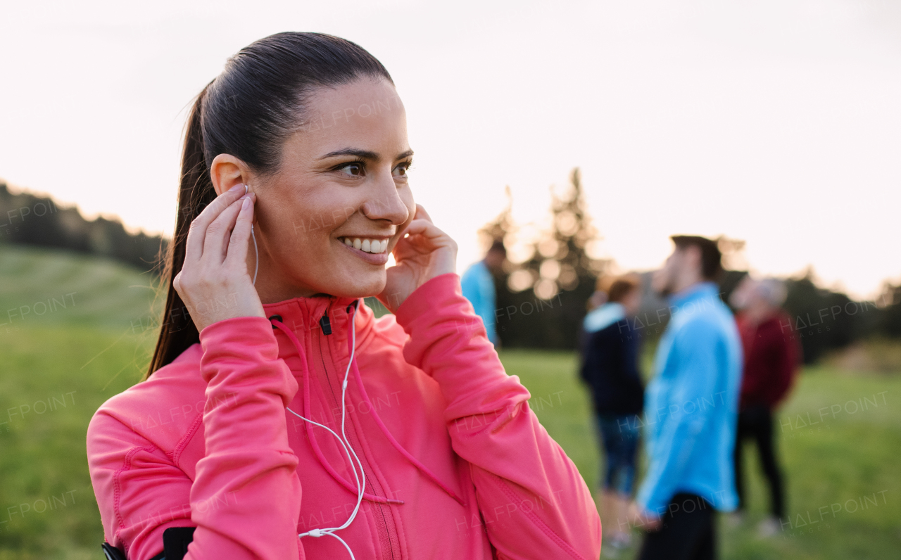 A portrait of young woman with earphones and group of people doing exercise in nature, resting.