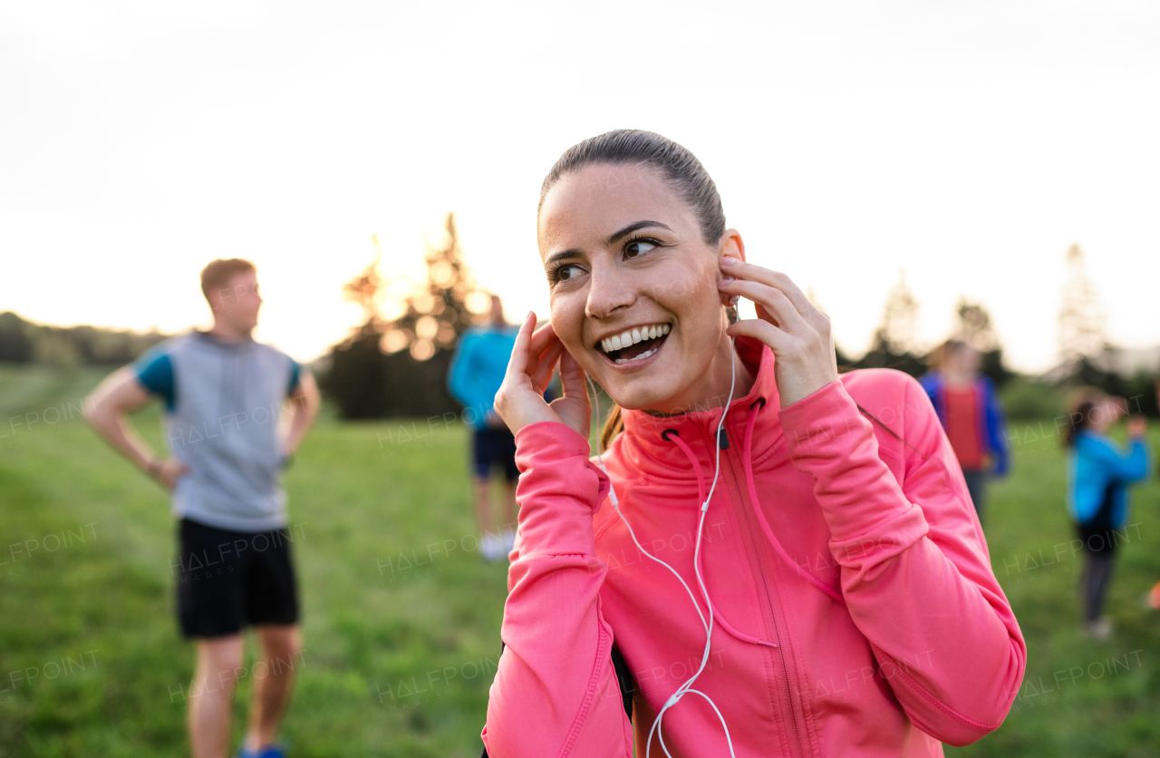 A portrait of young woman with earphones and group of people doing exercise in nature, resting.