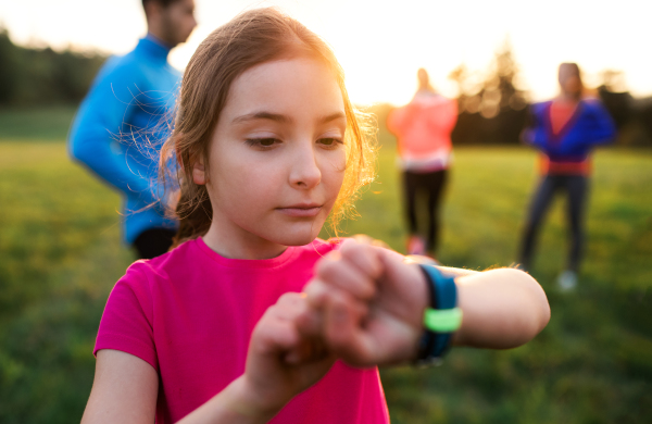 A portrait of small girl with large group of people doing exercise in nature, using smartwatch.