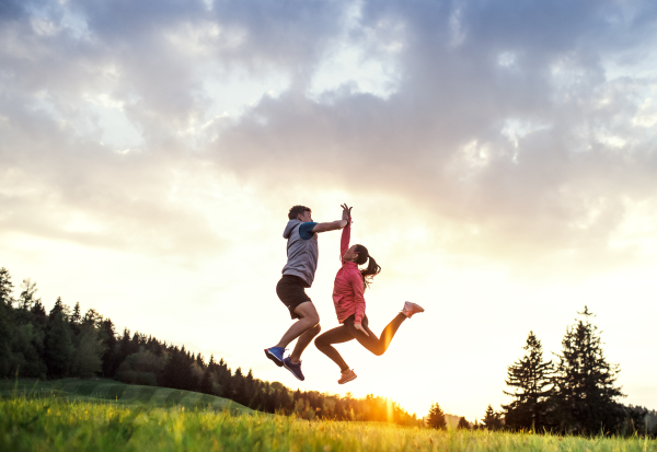 An active and fit senior couple jumping after doing exercise in nature at sunset, dusk sky in the background.