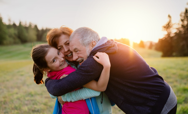 An active senior couple with cheerful granddaughter standing in nature at sunset.