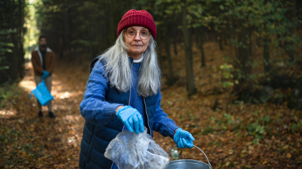 A senior woman volunteer looking at camera and cleaning up forest from waste, community service concept