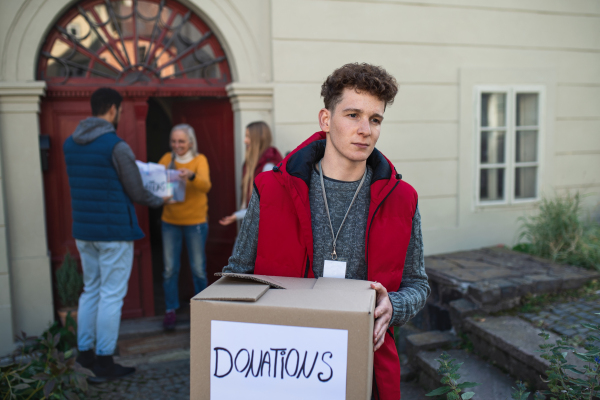 A group of young volunteers giving donation box to senior woman standing in door, social care concept