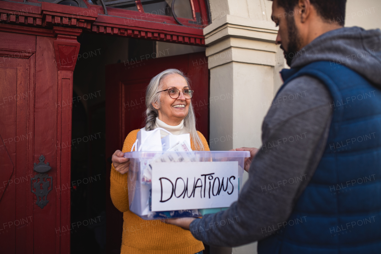 A young volunteer giving donation box to senior woman standing in doorway, social and charity concept