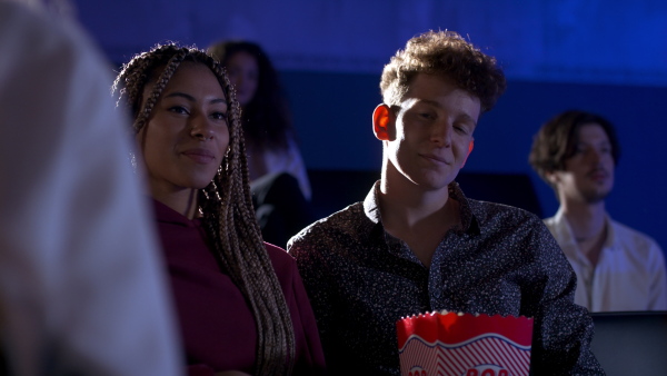 Cheerful young couple eating popcorn while watching movie in cinema, hugging and looking at camera.