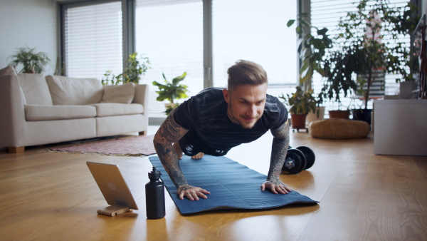 Front view portrait of young man with tablet doing push-ups workout exercise indoors at home.