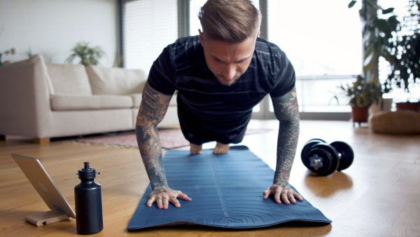 Front view portrait of young man with tablet doing push-ups workout exercise indoors at home.