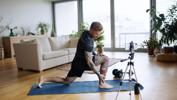 Young man trainer doing online workout exercise indoors at home, using camera.