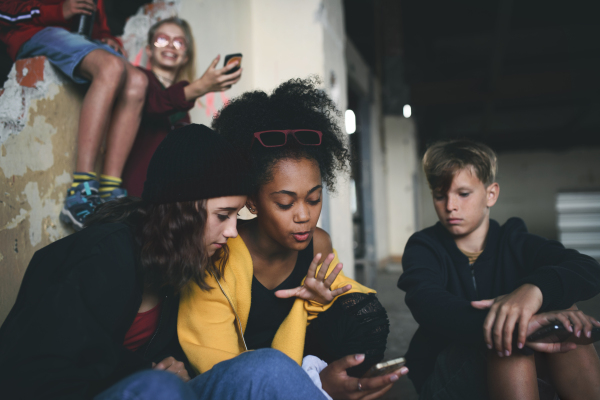 Front view of group of teenagers gang sitting indoors in abandoned building, using smartphones.