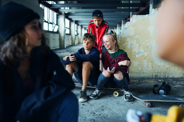 Front view of group of teenagers gang sitting indoors in abandoned building, using smartphones.