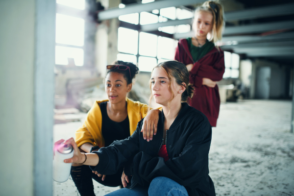 Group of teenagers girl gang standing indoors in abandoned building, using spray paint on wall.