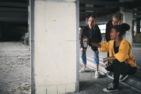 Group of teenagers girl gang standing indoors in abandoned building, using spray paint on wall.