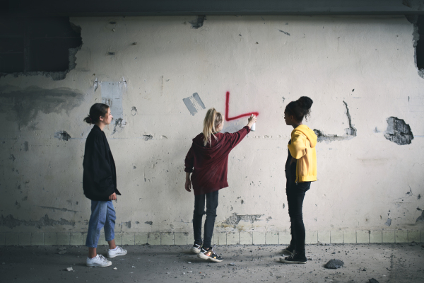 Rear view of group of teenagers girl gang standing indoors in abandoned building, using spray paint on wall.