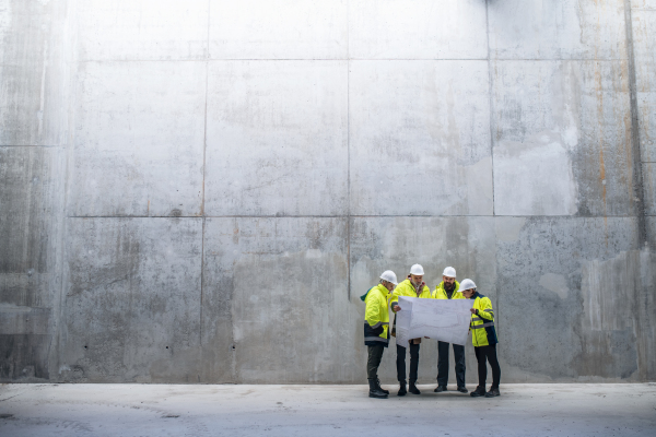 A group of engineers standing against concrete wall on construction site, holding blueprints. Copy space.