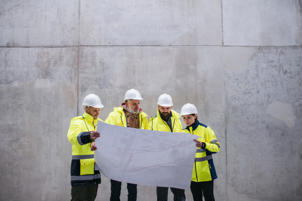 A group of engineers standing against concrete wall on construction site, holding blueprints. Copy space.