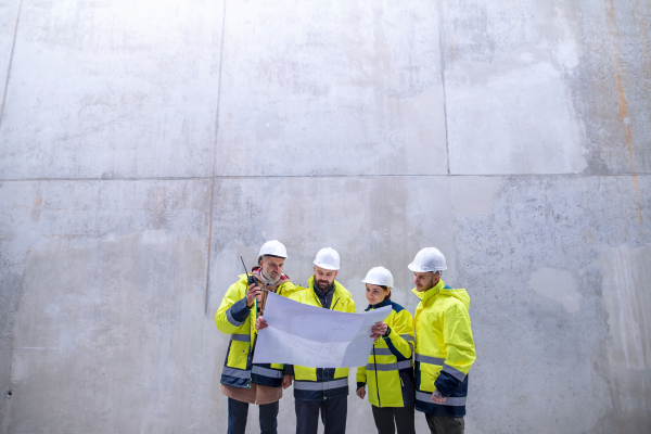 A group of engineers standing against concrete wall on construction site, holding blueprints. Copy space.