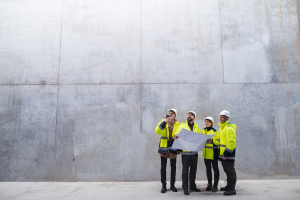 A group of engineers standing against concrete wall on construction site, holding blueprints. Copy space.