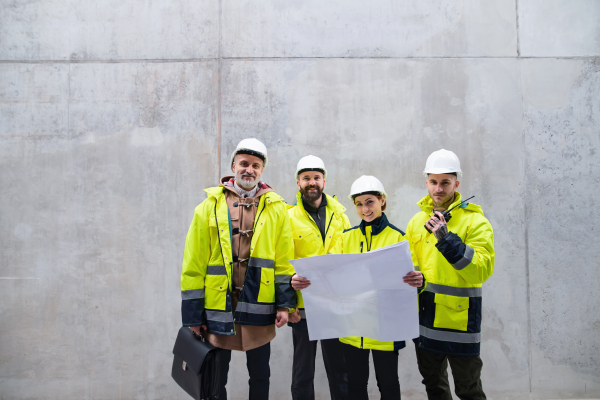 A group of engineers standing against concrete wall on construction site, holding blueprints. Copy space.