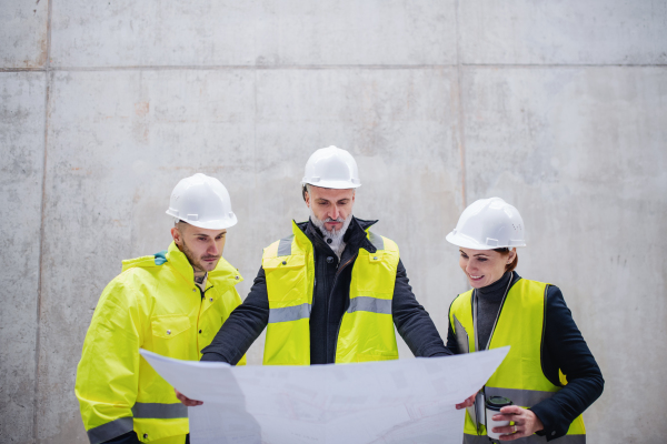 A group of engineers standing against concrete wall on construction site, holding blueprints.