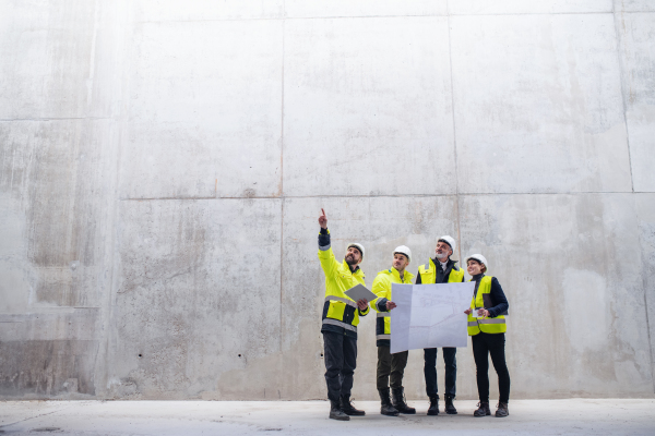 A group of engineers standing against concrete wall on construction site, holding blueprints. Copy space.