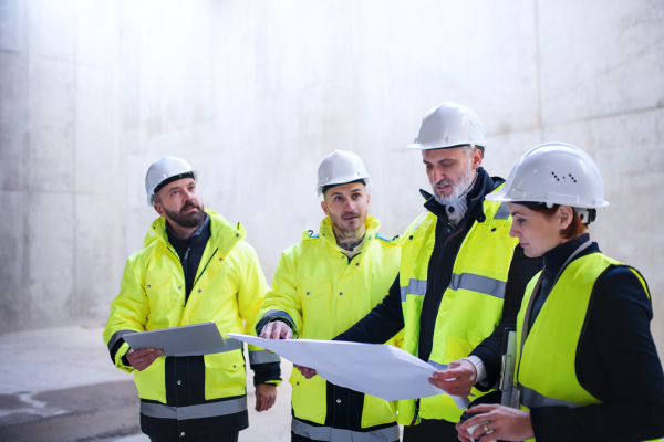 A group of engineers standing against concrete wall on construction site, holding blueprints. Copy space.