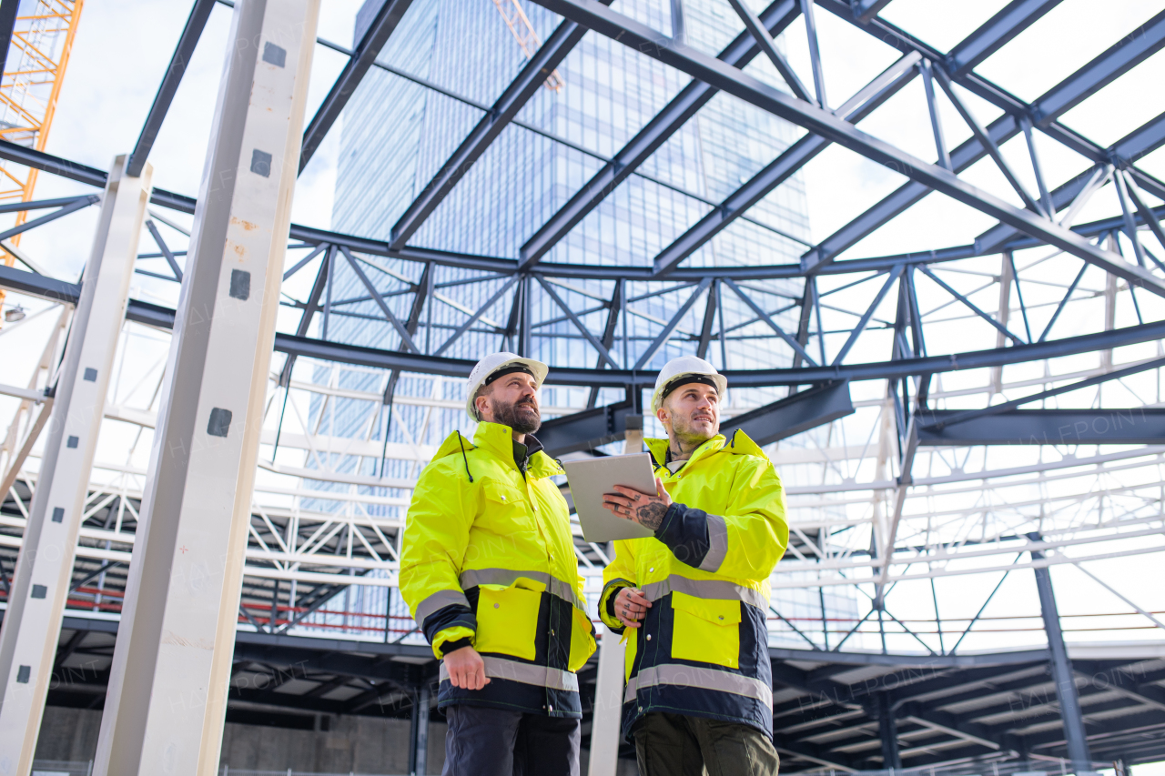 Front view of men engineers standing outdoors on construction site, using tablet.