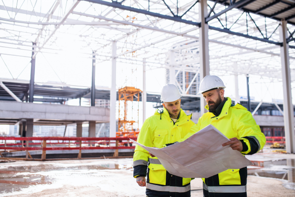 Front view of men engineers standing outdoors on construction site, holding blueprints.