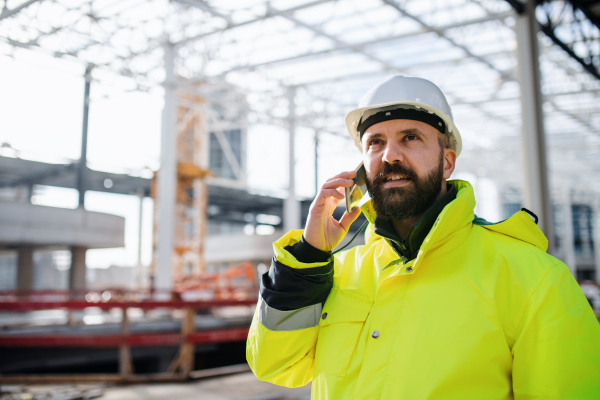 Mature man engineer standing on construction site, using smartphone.