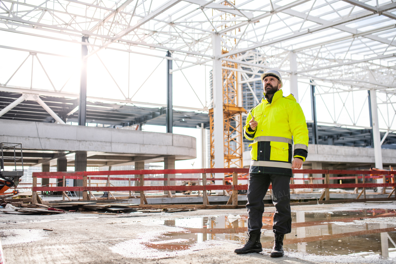 Mature man engineer standing on construction site, using smartphone.