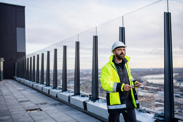 Mature man engineer walking on construction site, holding tablet with blueprints.