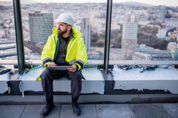 Mature man engineer sitting on construction site, holding tablet with blueprints.