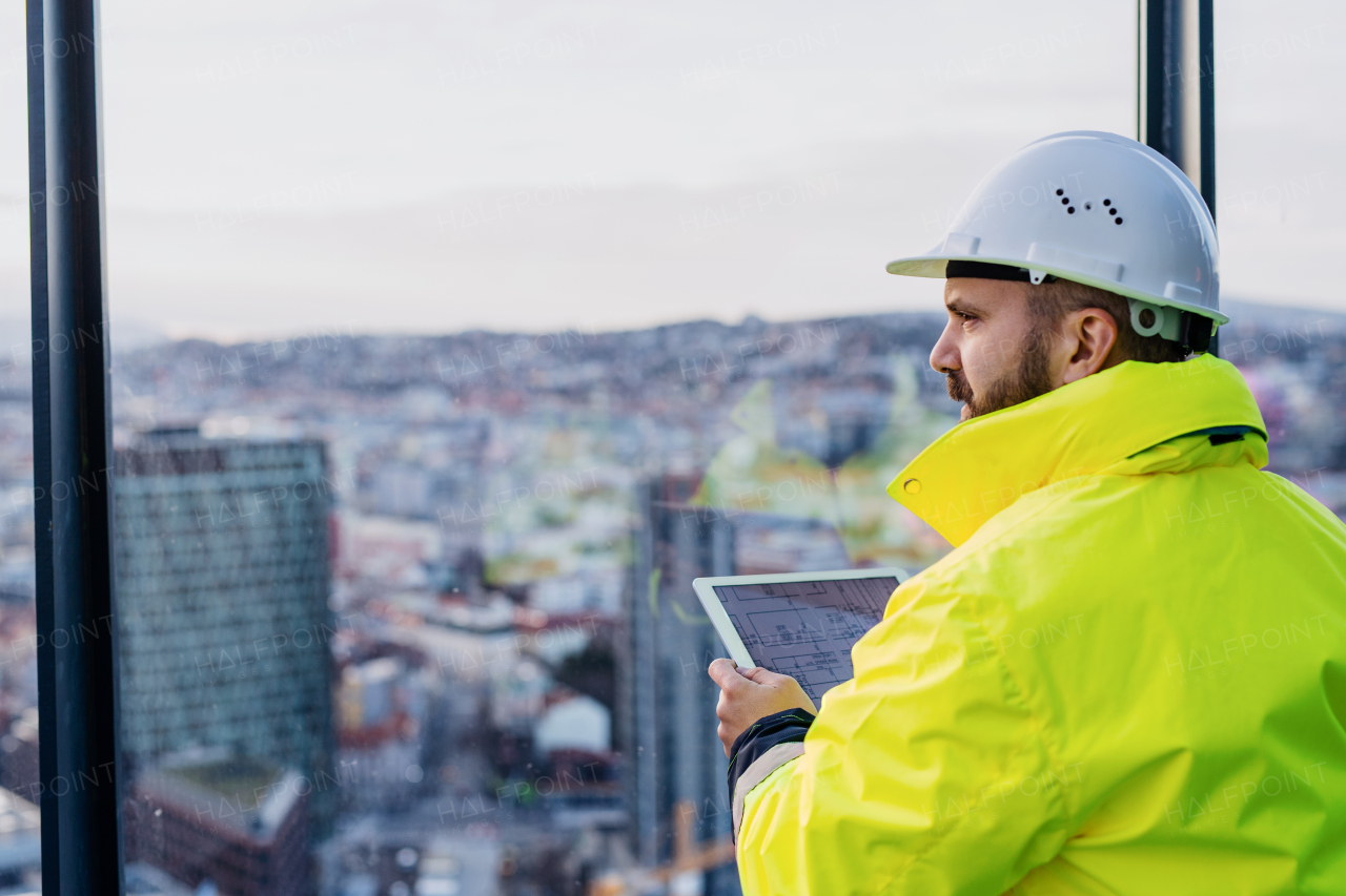 Rear view of man engineer standing on construction site, holding tablet with blueprints. Copy space.