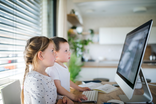 Small girl and boy sitting at the table, using computer indoors at home. Corona virus and quarantine concept.