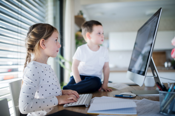 Small girl and boy sitting at the table, using computer indoors at home. Corona virus and quarantine concept.