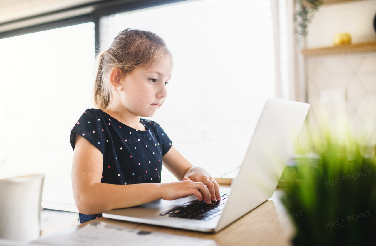 Small girl sitting at the table and using laptop indoors at home, Corona virus and quarantine concept.