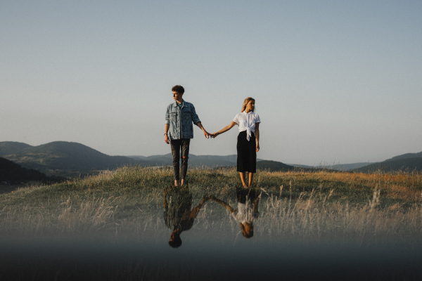 A portrait of young couple standing in nature in countryside, holding hands but looking away from each other.