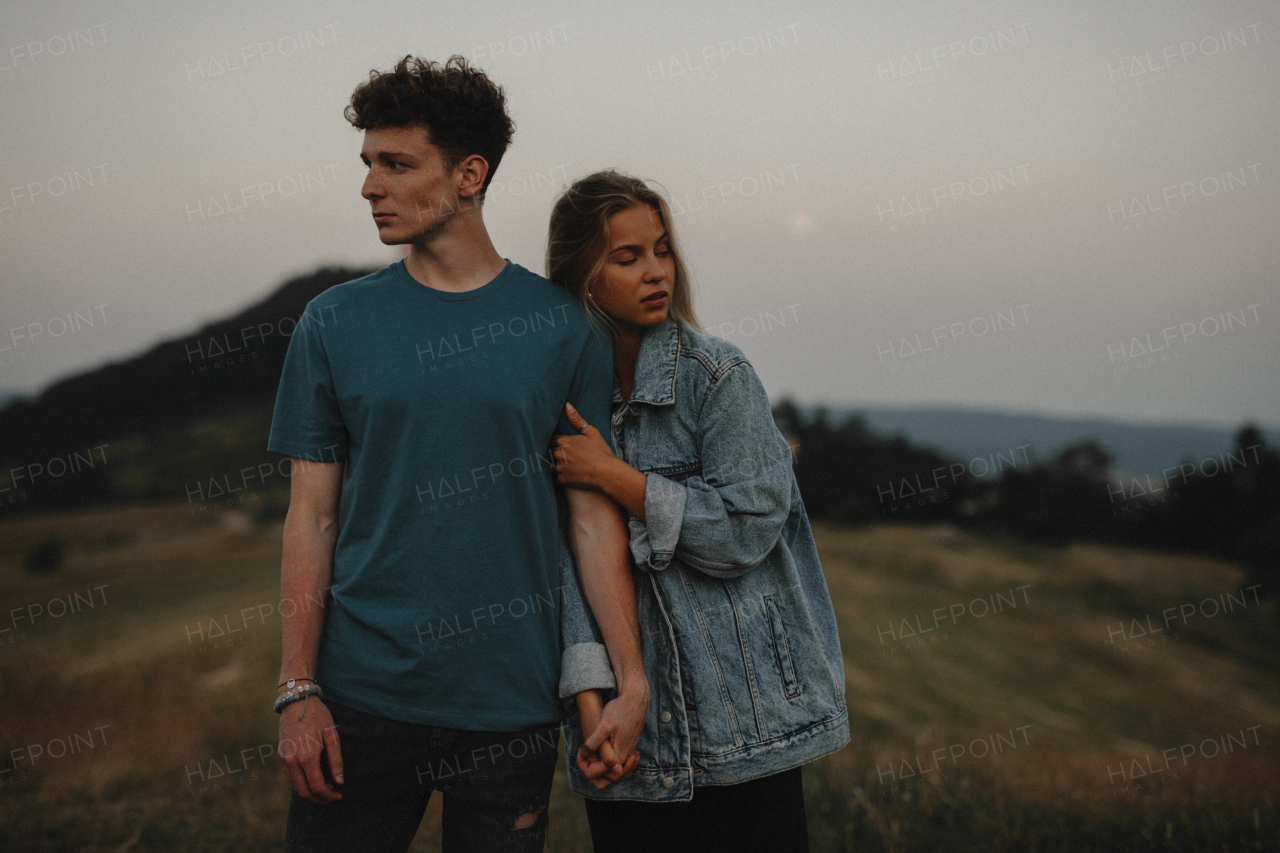 A portrait of young couple standing in nature in countryside, holding hands but looking away from each other.