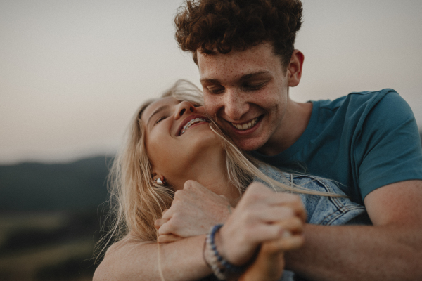 Side view of young couple on a walk in nature at sunset in countryside, hugging and having fun.