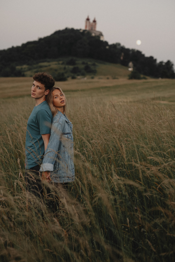 A young couple on a walk in nature at sunset in countryside, standing back to back and looking at camera.