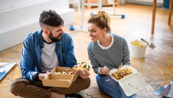 Mid adults couple eating lunch indoors at home, relocation, diy and food delivery concept.