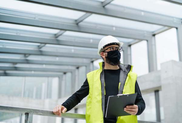 Portrait of worker with face mask, helmet and high visibility vest standing at the airport, holding clipboard.