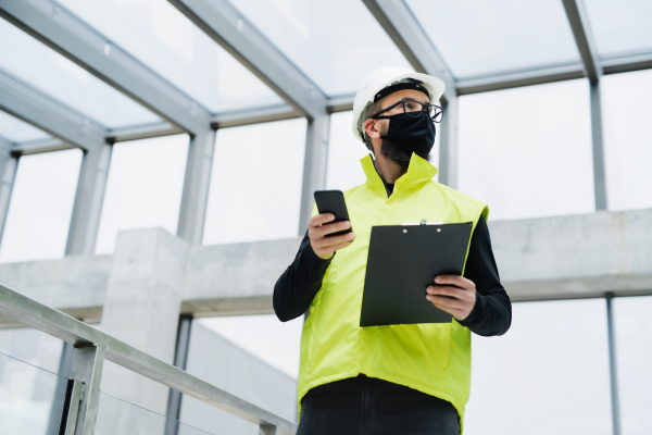 Portrait of worker with face mask, helmet and high visibility vest standing at the airport, holding smartphone.