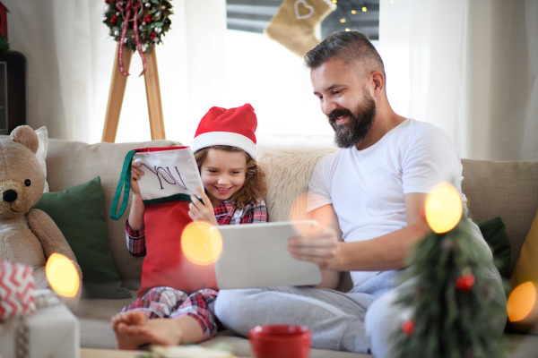 Portrait of father with small daughter indoors at home at Christmas, using tablet.