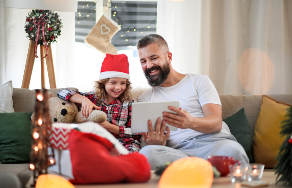 Portrait of father with small daughter indoors at home at Christmas, using tablet.