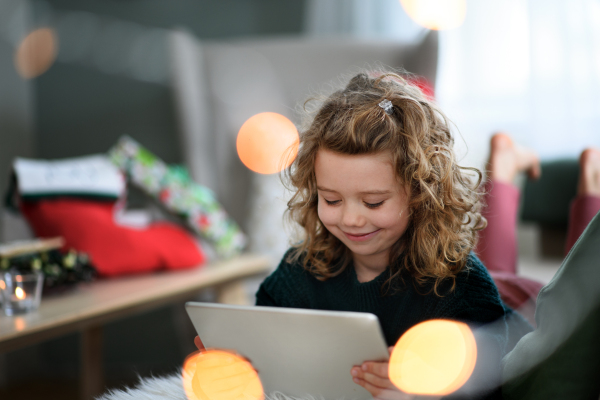 Portrait of happy small girl on bed indoors at home at Christmas, using tablet.