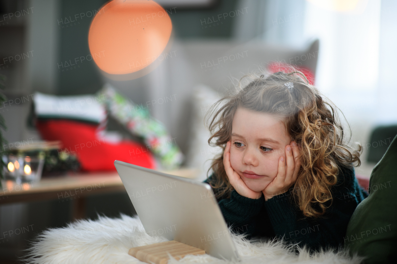 Portrait of sad small girl on bed indoors at home at Christmas, using tablet.