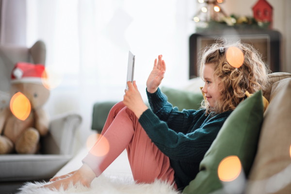 Small girl wth tablet sitting indoors at home at Christmas, having video call.