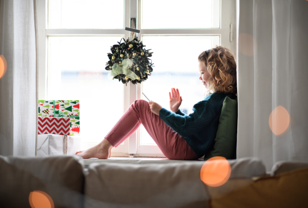 A small girl with tablet sitting indoors at home at Christmas, having video call.