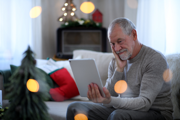 Portrait of sad and lonely senior man with tablet sitting at the table indoors at Christmas, solitude concept.