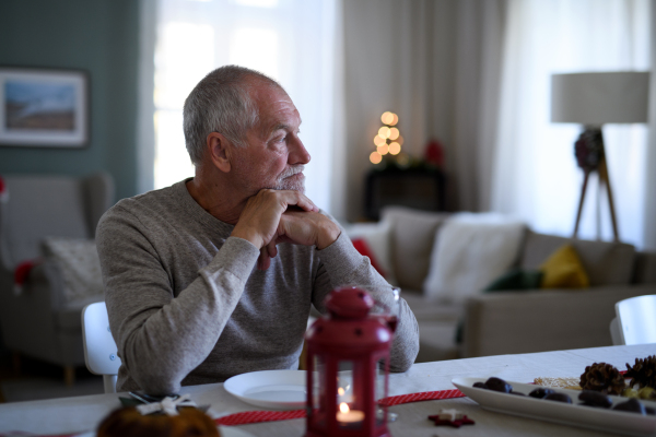 Portrait of lonely senior man sitting at the table indoors at Christmas, solitude concept.